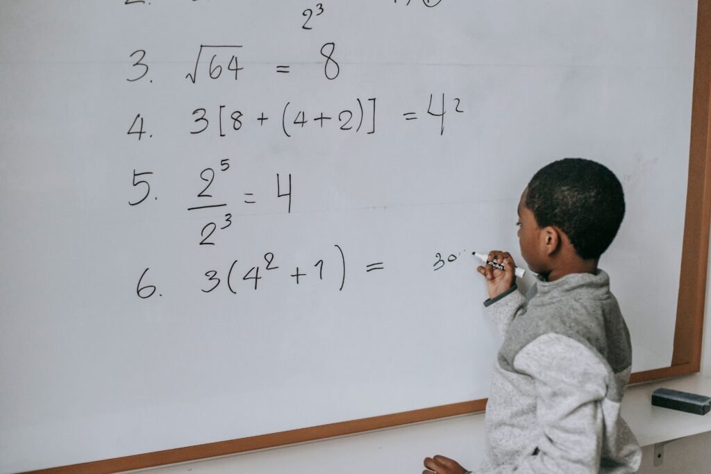 Side view of concentrated African American pupil standing near whiteboard in classroom and solving math examples during lesson at school