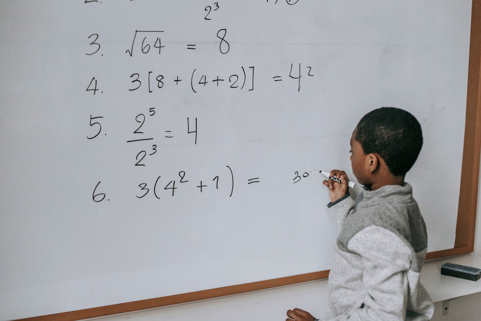 Side view of concentrated African American pupil standing near whiteboard in classroom and solving math examples during lesson at school