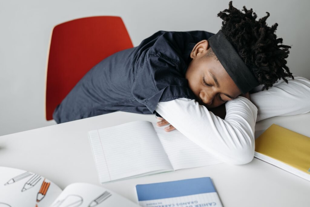 A young boy with dreadlocks asleep on his desk in a classroom setting, surrounded by notebooks.